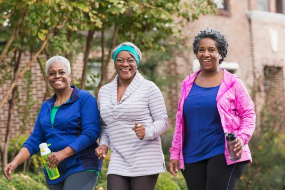 Un grupo de tres mujeres afroamericanas mayores haciendo ejercicio juntas, caminando por una acera en un vecindario residencial. Se están divirtiendo, sonriendo a la cámara.